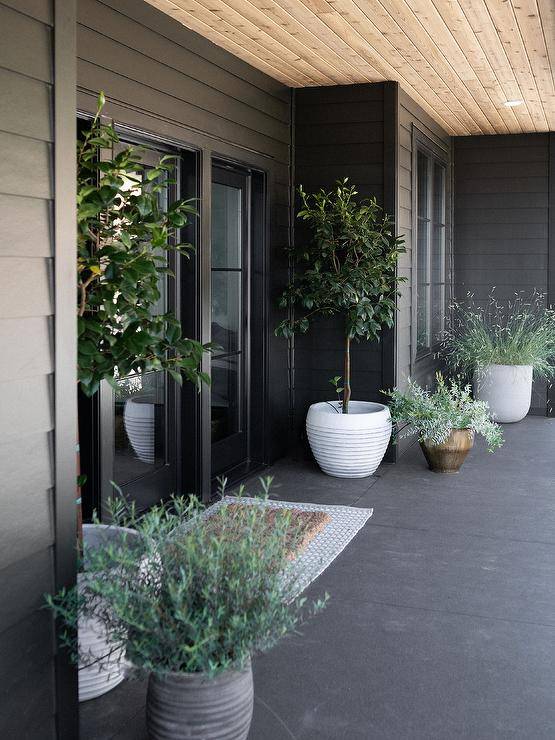 A black clapboard home is accented with black front doors flanked by potted plants placed on either side of layered rugs.