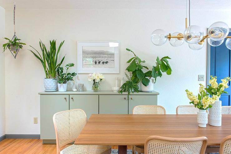 Transitional dining room features a vintage cane dining chairs at an oak table illuminated by glass and brass globe lights. A green buffet dresser displays house plants for a welcoming and natural finish.