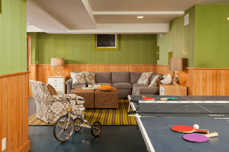 Cool retro basement game room design with contemporary ceiling with pot lights and fresh green grasscloth wallpaper, paired with unstained wood paneling on half walls. Ping pong table provides the fun while a roll-armed gray sectional sofa paired with cork cube ottomans function as perfect seating upon a yellow and brown stripes.