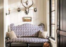 Skull mounts are fixed to vertical shiplap trim over a small landscape art piece hung above a purple upholstered settee topped with black check pillows placed on a dark stained wood floor. Natural light streams in from a sidelight located beside a black door.