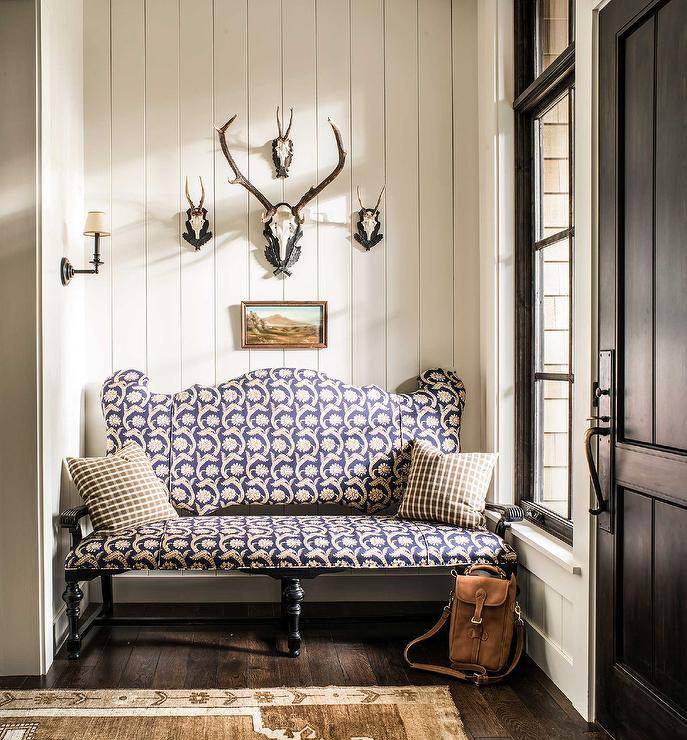 Skull mounts are fixed to vertical shiplap trim over a small landscape art piece hung above a purple upholstered settee topped with black check pillows placed on a dark stained wood floor. Natural light streams in from a sidelight located beside a black door.