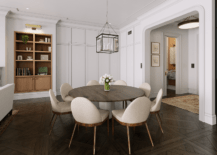 Dining space features linen and walnut dining chairs at a round walnut dining table on parquet wood floors, illuminated by a square lantern and brown built in shelves.