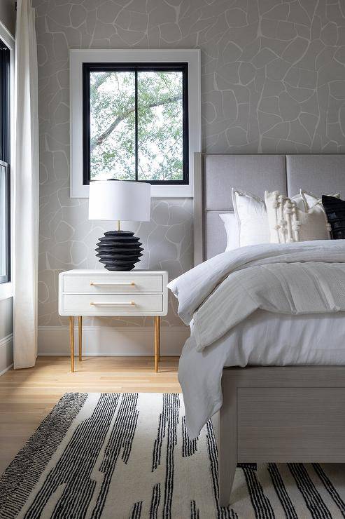 Bedroom features a light gray wood and fabric bed atop a black and white rug and a black lamp that illuminates a white and gold nighstand on white and gray wallpaper.