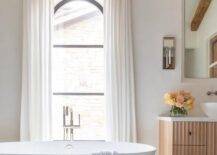 Bathroom features an oval bathtub on marble hexagon tiles under an arched window with white curtains.