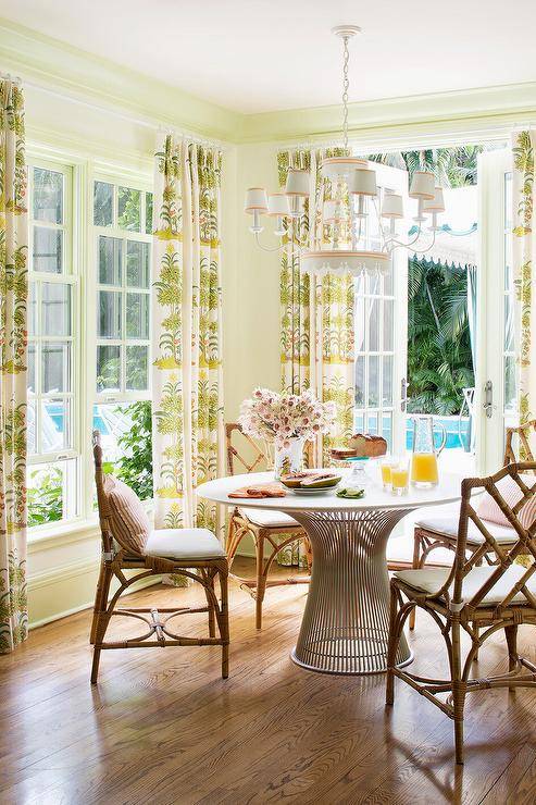 Positioned on a wood floor in a welcoming dining room, Platner Dining Table is surrounded by brown bamboo rattan dining chairs. Window are covered in white and green curtains.