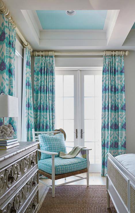 Turquoise blue ikat curtains hang in front of a glass paneled French doors positioned behind a white accent chair fitted with a rush seat topped with blue cushions. A gray wood dresser sits beneath a turquoise blue coffered ceiling and behind a bound seagrass rug.
