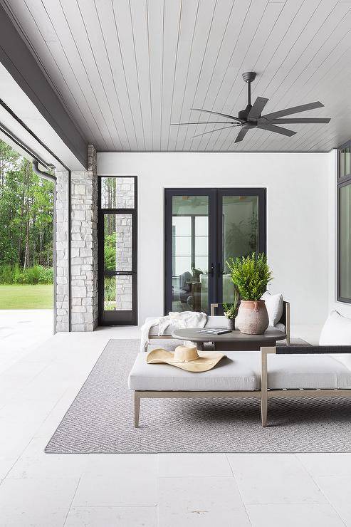 A gray diamond jute rug is placed on a covered patio beneath a gray coffee flanked by gray teak and rope loungers cooled by a black modern ceiling fan fixed to a gray beadboard ceiling.