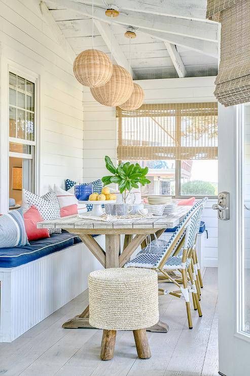 Beautiful enclosed patio boasts shiplap walls and wicker lanterns hung over a plank trestle dining table seating blue French bistro chairs facing a beadboard dining bench topped with blue cushions.