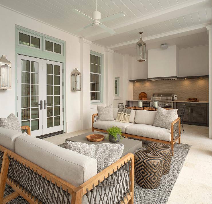Small enclosed patio features wood and gray rope outdoor sofas that face a gray outdoor coffee table with brown stools atop a gray rug and an outdoor kitchen with brown cabinets lit by a nickel lantern.