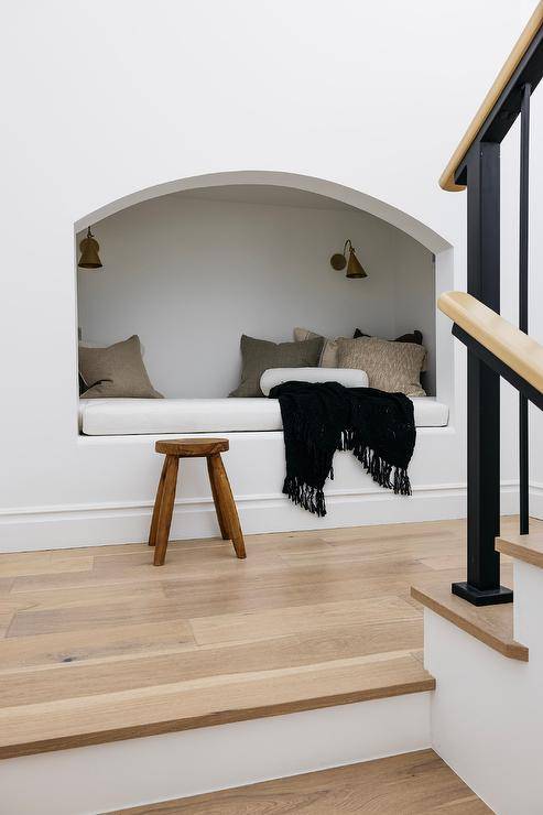Brass sconces illuminated a staircase with built in reading nook accented with tan pillows and a black throw blanket and a wooden stool.