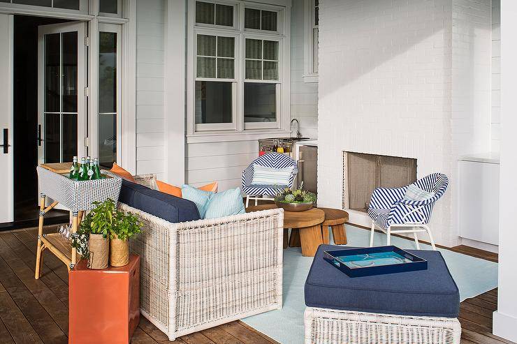 Covered patio featuring a bar sink beside a white brick fireplace furnished with blue herringbone outdoor chairs, nesting coffee tables and white wicker sofa seating. The bar sink includes a stainless steel mini fridge bringing a small kitchen space outdoor perfect for entertaining guests.