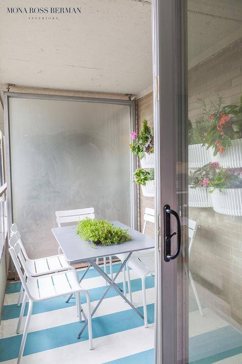 A sliding glass door opens to a small enclosed patio filled with a gray x base dining table and white chairs placed atop a white and turquoise blue striped rug facing a brick wall lined with stacked flower planters.