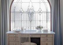 Gray curtains hang on either side of an arched stained glass window located behind a hand scraped oak desk placed on marble floor tiles and matched with an oak desk chair.