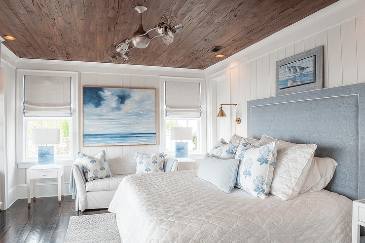 Under a rustic stained plank ceiling, this restful white and blue cottage bedroom boasts an art piece placed atop a blue linen headboard positioned in front of a white shiplap wall and behind a bed dressed in white bedding accented with white and blue pillows. The bed sits between white nightstands lit by brass swing arm sconces, while a white slipcovered sofa is placed beneath a large sea art piece . The art hangs between windows covered in gray grosgrain roman shades hung above white end tables lit by blue lamps.
