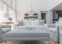 Lovely light gray and blue living room features a white sofa accented with blue pillows complementing a blue ottoman coffee table finished with lucite legs and placed on a gray rug between facing gray slipcovered wingback chairs.