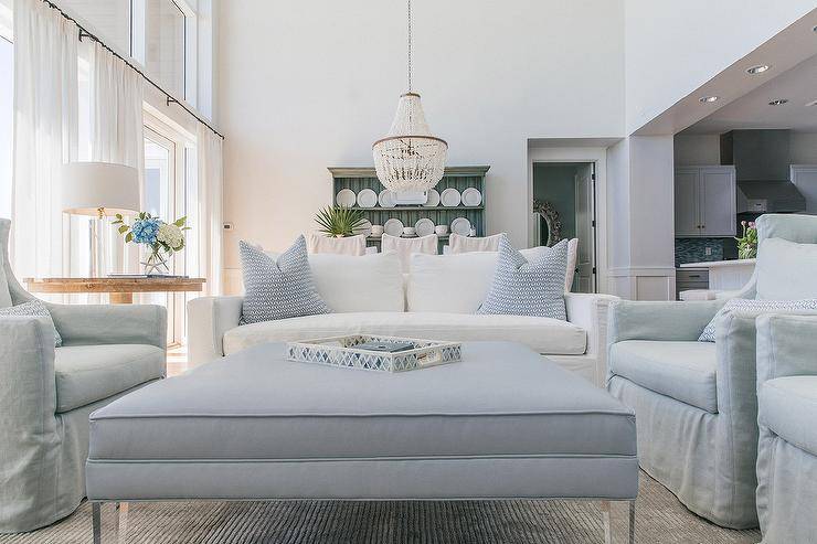 Lovely light gray and blue living room features a white sofa accented with blue pillows complementing a blue ottoman coffee table finished with lucite legs and placed on a gray rug between facing gray slipcovered wingback chairs.