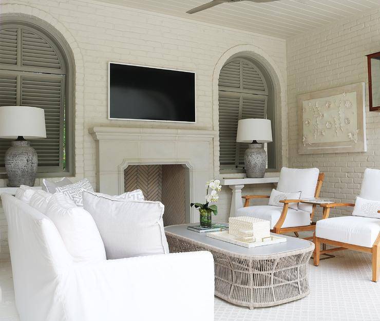 White brick frames arched windows covered in gray shutters and positioned flanking a flat panel television mounted over a limestone fireplace. Distressed gray lamps sit on white console tables beneath the windows. Beneath a plank ceiling, a white slipcovered sofa topped with gray sofa pillows sits on light gray lattice rug at a styled light gray oval wicker coffee table.