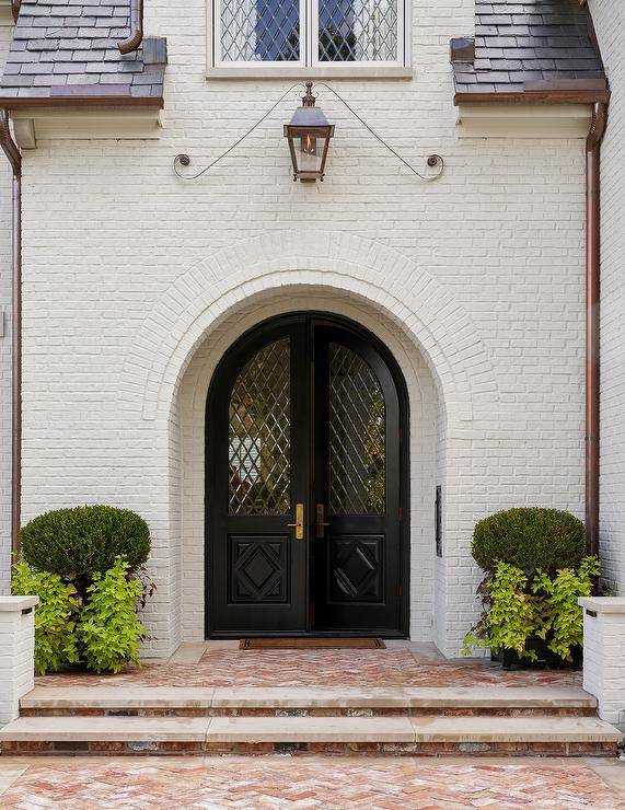 A white brick French chateau features black lattice front doors lit by a lantern and red brick herringbone pavers.