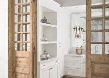 Gorgeous vintage folding pantry doors are accented with glass panels and open to a white pantry boasting a wide plank wood floor and white cabinets with polished nickel knobs.