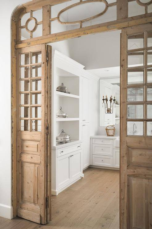Gorgeous vintage folding pantry doors are accented with glass panels and open to a white pantry boasting a wide plank wood floor and white cabinets with polished nickel knobs.