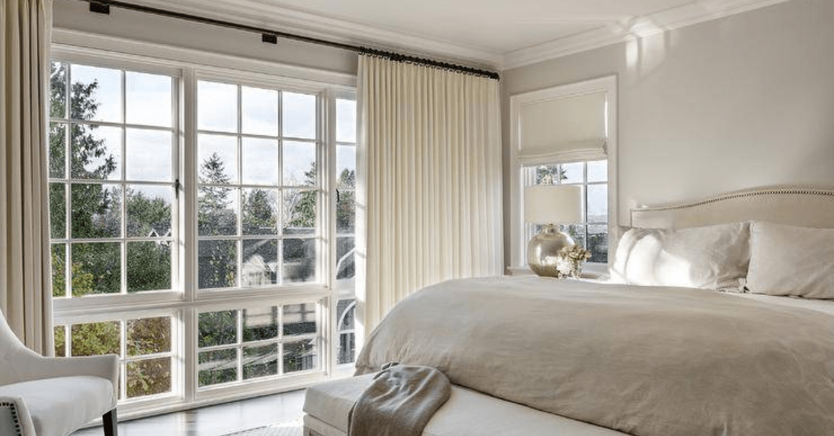 Cozy bedroom features a light tan camelback headboard lit by a mercury glass lamp, a tan bench at tthe foot of the bed and cream curtains.