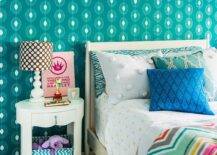 Teal teen girl's bedroom features an ivory bed dressed in green star sheets and a colorful chevron duvet accented with a turquoise trellis print pillow, a blue diamond print pillow, and a teal zebra print body pillow all sat in front of a teal wallpapered wall. Beside the bed, a Bungalow 5 Brigitte 1-Drawer side table holding a teal chevron basket on the bottom and topped with a brown polka dotted four gourd white lamp and a pink canvas print is sat on a striped apple green area rug.