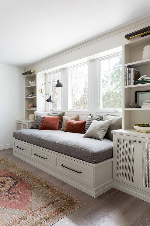 A white built-in day bed is fitted with drawers donning oil rubbed bronze pulls and is topped with a gray cushion positioned under a row of windows. The daybed is flanked by white built-in shelves lit by brass picture lights and mounted over cabinets.