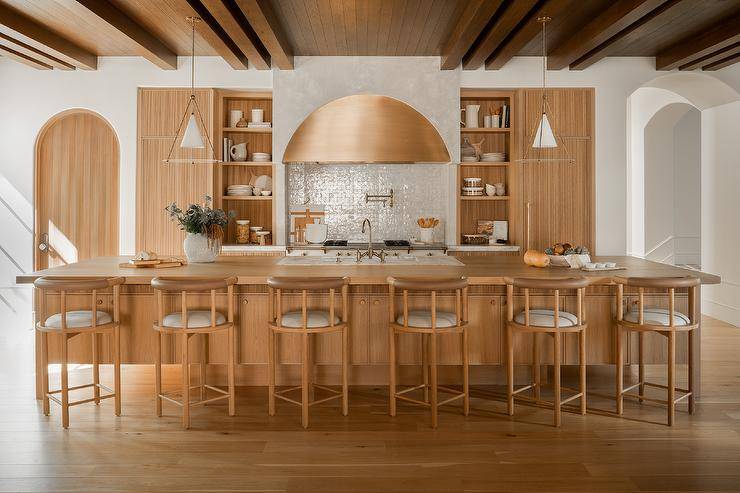 Spacious kitchen features wood and leather barrel back stools at a reeded center island with wood countertop lit by conical pendants.
