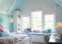 A gorgeous chandelier hangs from a blue vaulted ceiling in a pink and blue girl's bedroom furnished with a white wood bed accented with pink and blue suzani style bedding. The bed sits on a pink trellis rug facing a blue desk seating a white faux fur chair. A white built-in window seat is finished with a blue cushion flanked by facing white built-in bookcases, while windows frame by white shiplap are dressed in white roman shades.
