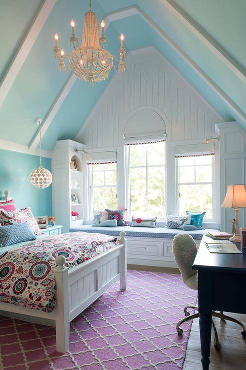 A gorgeous chandelier hangs from a blue vaulted ceiling in a pink and blue girl's bedroom furnished with a white wood bed accented with pink and blue suzani style bedding. The bed sits on a pink trellis rug facing a blue desk seating a white faux fur chair. A white built-in window seat is finished with a blue cushion flanked by facing white built-in bookcases, while windows frame by white shiplap are dressed in white roman shades.