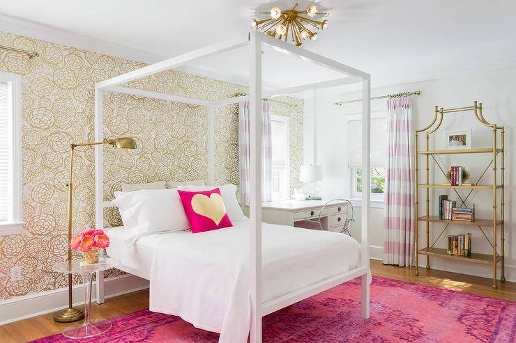 Pink and gold teenage girl's bedroom features a brass sputnik flush mount, Jonathan Adler Sputnik 12 Light, placed over a white canopy bed dressed in white bedding and a pink and gold metallic heart pillow lining an accent wall clad in gold metallic wallpaper, Oh Joy Petal Pusher Wallpaper, atop a hot pink overdyed rug. A white canopy bed is flanked by an acrylic tulip table and a brass floor pharmacy lamp to the left and a white desk with Ghost Chair to the right. Chic girl's bedroom boasts a gold metal bamboo etagere placed next to windows dressed in pink striped curtains layered over white roman shades.