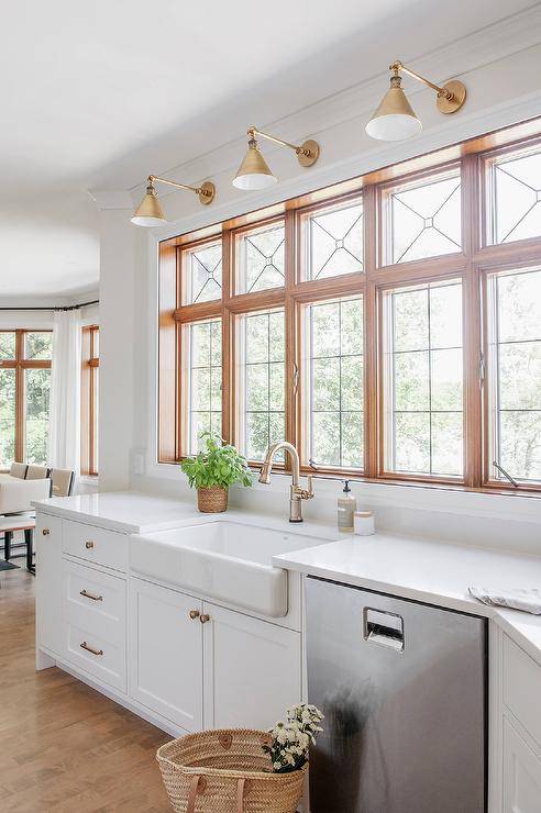 Brass hardware complements white kitchen cabinets holding a farmhouse sink beneath a brass gooseneck faucet mounted under leaded glass windows illuminated by Boston Functional Library Lights.