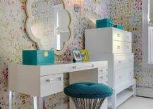 Chic teen girl's bedroom features walls clad in Kandy Brit Pop Wallpaper lined with a white vanity adorned with lucite pulls and lucite legs paired with a turquoise Greek key stool under a cream scalloped mirror, Made Goods Fiona Mirror, next to a Bungalow 5 Pandora Tall 4 Drawer.