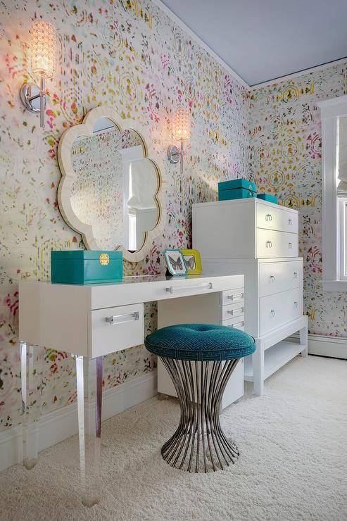 Chic teen girl's bedroom features walls clad in Kandy Brit Pop Wallpaper lined with a white vanity adorned with lucite pulls and lucite legs paired with a turquoise Greek key stool under a cream scalloped mirror, Made Goods Fiona Mirror, next to a Bungalow 5 Pandora Tall 4 Drawer.