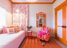 Lovely pink and orange girl's bedroom features a white Jenny Lind bed placed beneath a window covered in white plantation shutters dressed in light pink bedding topped with pink and orange pillows. White and pink curtains hang behind the bed under a ceiling fitted with orange crown moldings. An orange and pink accent chair sits on a pink and orange rug in front of an orange mirror and beside a red quatrefoil accent table. The room is completed with stunning orange and light pink double closet doors with oil rubbed bronze knobs and a pale pink painted ceiling.