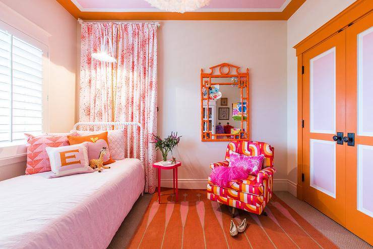 Lovely pink and orange girl's bedroom features a white Jenny Lind bed placed beneath a window covered in white plantation shutters dressed in light pink bedding topped with pink and orange pillows. White and pink curtains hang behind the bed under a ceiling fitted with orange crown moldings. An orange and pink accent chair sits on a pink and orange rug in front of an orange mirror and beside a red quatrefoil accent table. The room is completed with stunning orange and light pink double closet doors with oil rubbed bronze knobs and a pale pink painted ceiling.