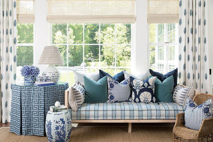 Paisley block print curtains hang from a window covered in bamboo roman shades hung above a cane day bed accented with a blue stripe cushion. The daybed sits on a bound jute rug beside an octagon skirted end table lit by a white and blue tiled chevron lamp.