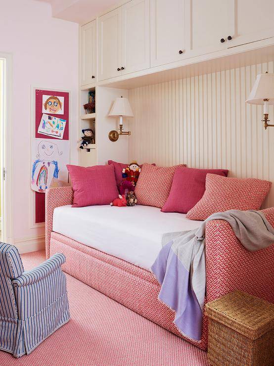 Comfy, cozy and bright pink geometric daybed dressed with matching accent pillows and a lavender fringy throw blanket against a white beadboard wall under built-in storage cabinets lit by white shade swing arm sconces.