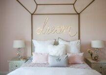 A 'dream' marquee light is mounted to a pink wall behind a gold canopy bed dressed in white and pink bedding topped with layered white and gray shams placed behind pink and gray pillows. The bed sits between black and white bone inlay nightstands lit by stacked glass gem lamps.