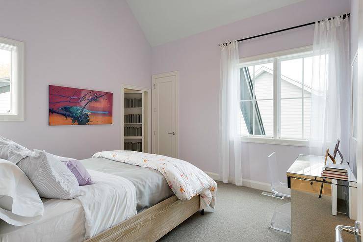 In this beautiful lilac girl's bedroom, a stained oak bed is dressed in purple and gray bedding and faces a window framed by a lilac wall and covered in sheer white curtains. To the side of the bed, a lucite z-chair sits at a mirrored desk placed on gray carpeting.
