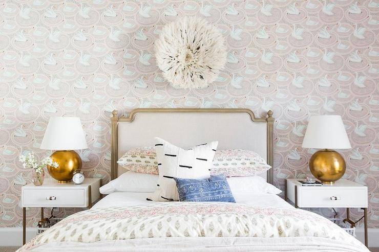 A juju hat decorates a chic girl's bedroom above a gray French bed with green and pink block print bedding complimenting swan wallpaper. Two gold lamps adorn white nighstands adding a sophisticated appeal to the girl's bedroom space.