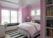 Gorgeous white and pink girl's bedroom is lit by an Ikea PS Maskros Pendant hung over a black and white striped rug place beneath a white upholstered bed paired with a white tufted headboard accenting pink and white bedding. The bed is topped with black and gold pillows layered in front of black and white check shams, and beneath a window framed by a pink wall. A white built-in window seat is fitted with storage drawers and a black seat cushion, while a white windsor chair with a black cushion is positioned at a two tone desk.