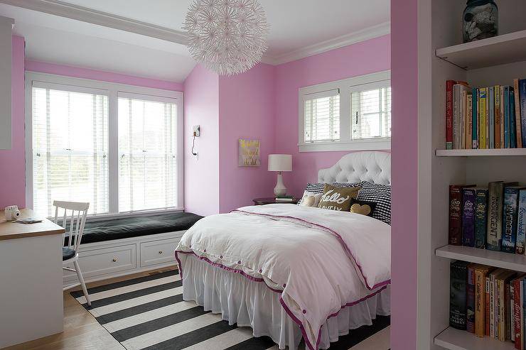 Gorgeous white and pink girl's bedroom is lit by an Ikea PS Maskros Pendant hung over a black and white striped rug place beneath a white upholstered bed paired with a white tufted headboard accenting pink and white bedding. The bed is topped with black and gold pillows layered in front of black and white check shams, and beneath a window framed by a pink wall. A white built-in window seat is fitted with storage drawers and a black seat cushion, while a white windsor chair with a black cushion is positioned at a two tone desk.