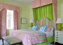 Cheery and fun pink, green and turquoise girl's bedroom with pale green walls and pink damask drapes and dramatic damask canopy behind a curvy striped headboard with matching striped ottoman at foot of bed. Beautiful white four drawer nightstands with green gourd style lamps and brass hardware flank bright pink and green girls bedding including blue geometric pillow and pink damask quilt. A pastel colored floral rug complete this dream girls bedroom.