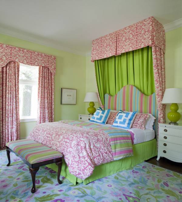 Cheery and fun pink, green and turquoise girl's bedroom with pale green walls and pink damask drapes and dramatic damask canopy behind a curvy striped headboard with matching striped ottoman at foot of bed. Beautiful white four drawer nightstands with green gourd style lamps and brass hardware flank bright pink and green girls bedding including blue geometric pillow and pink damask quilt. A pastel colored floral rug complete this dream girls bedroom.
