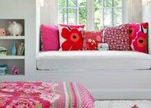 Tween girl's bedroom features a a white built in window seat topped with a white seat cushion accented with pink and red floral pillows placed in front of a dormer window. Beside the bench, recessed bookshelves face a gray linen bed placed on a white sheepskin rug and dressed in a red roses blanket.