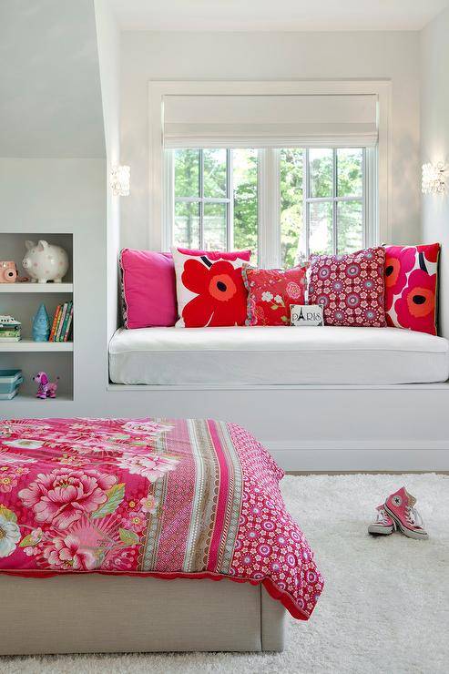 Tween girl's bedroom features a a white built in window seat topped with a white seat cushion accented with pink and red floral pillows placed in front of a dormer window. Beside the bench, recessed bookshelves face a gray linen bed placed on a white sheepskin rug and dressed in a red roses blanket.