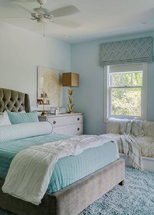 Luxurious aqua and gray girl's bedroom is furnished with a gray velvet tufted bed accented with aqua blue bedding topped with a large aqua and white bolster pillow. The bed sits on a blue shag rug beside a light gray French nightstand lit by a gold tulip lamp placed in front of a gold and white art piece hung from a soft blue wall. A white sheepskin couch is positioned beneath a window covered with a blue trellis roman shade.