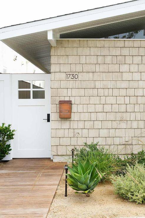 A tan shingled shaker siding home features a copper mailbox and a teak walkway leading to a white door.