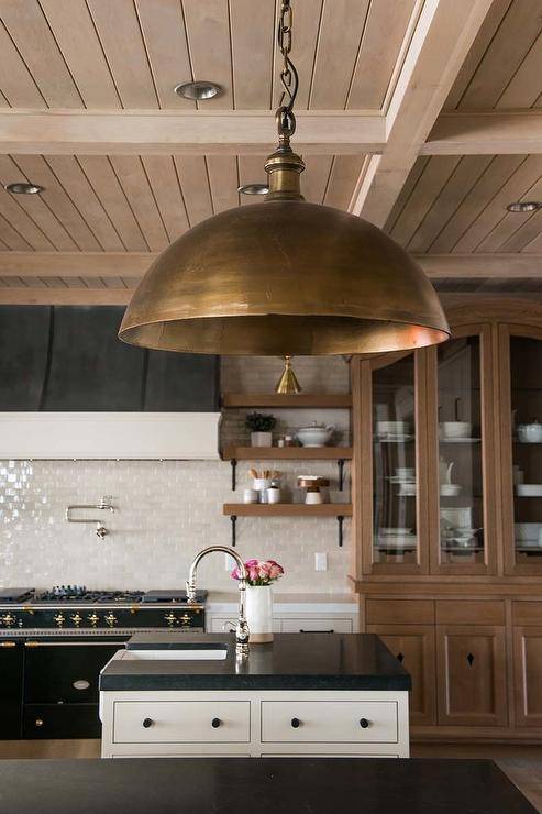 Lacanche range in a cottage kitchen designed with ivory crackled subway tiles, a polished nickel swivel arm pot filler, and a black and gold French range hood. Rustic wooden ceiling complements wood finishings and cooktop shelves for a rustic appeal.